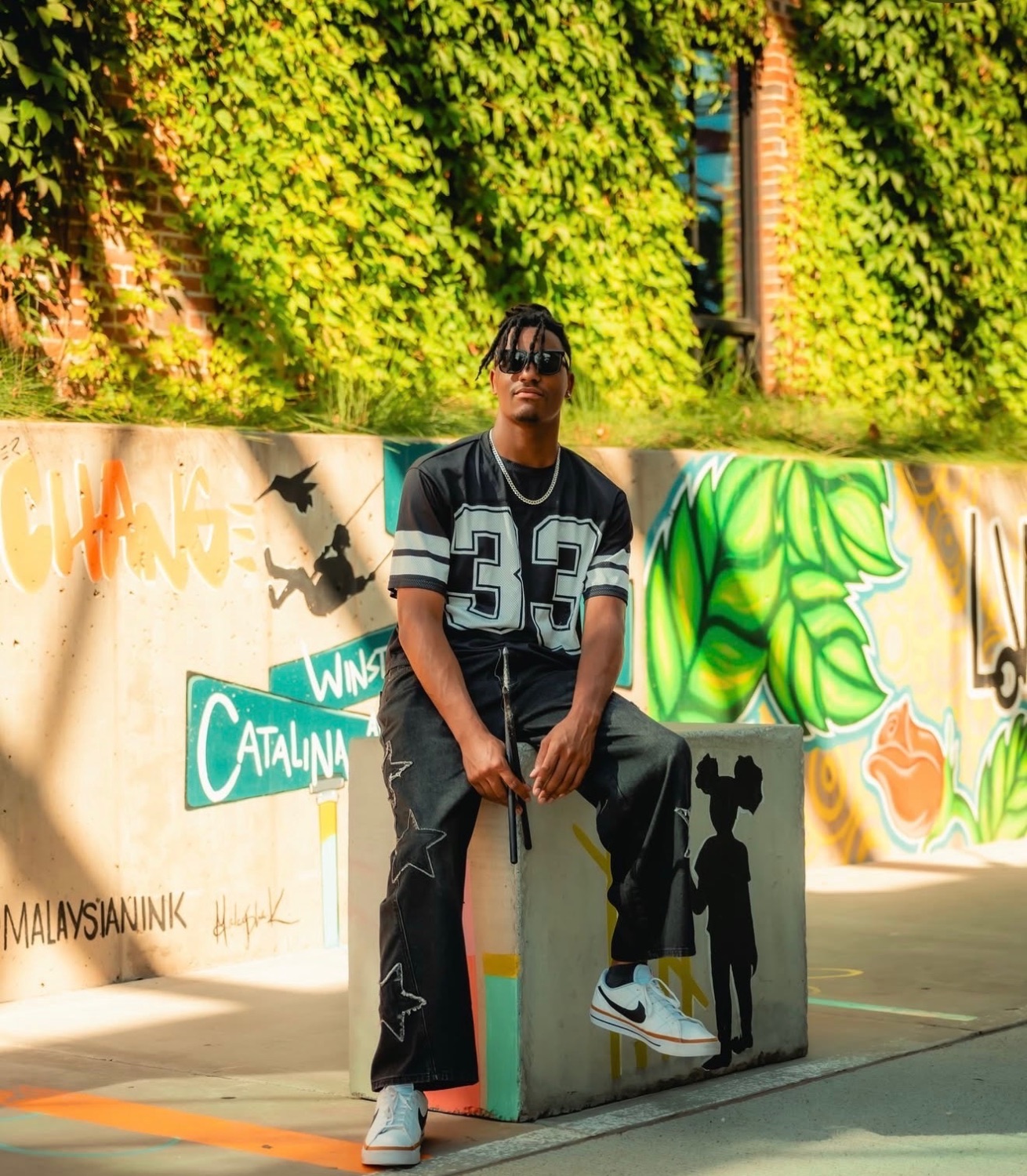 Jaywill Williams seated on a concrete block in front of a graffiti mural, wearing a number 33 jersey and sunglasses