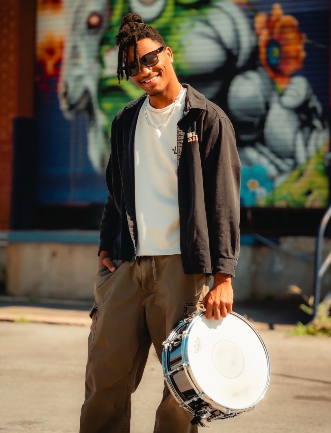 Jaywill Williams standing on a Charlotte street holding a snare drum, wearing sunglasses and a black jacket
